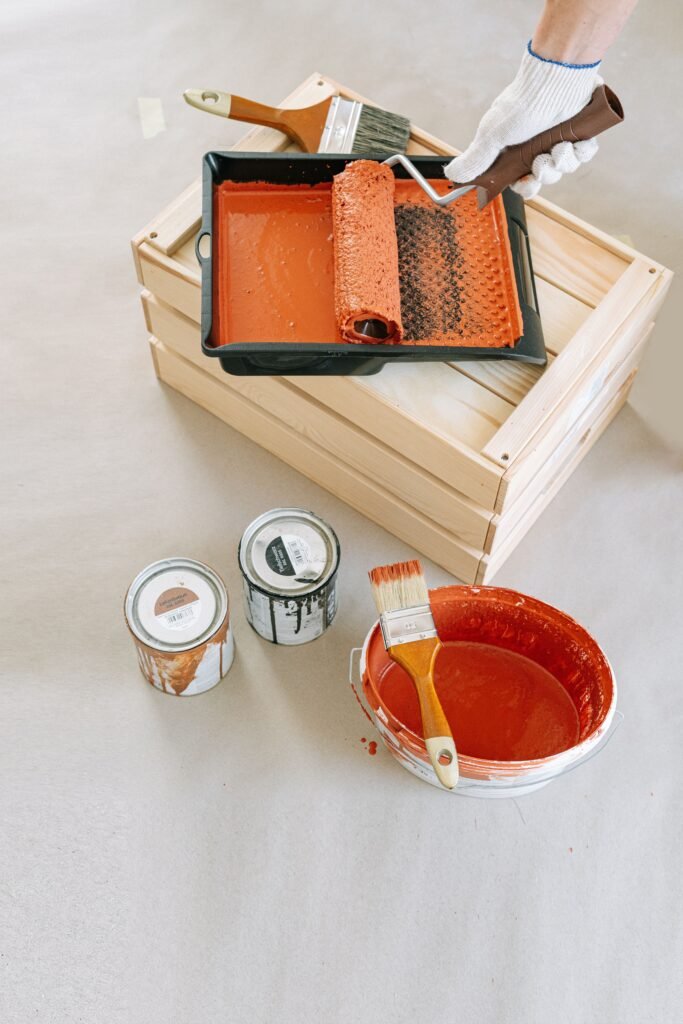 Flat lay of painting supplies on wooden crate, including roller, brushes, and paint cans.